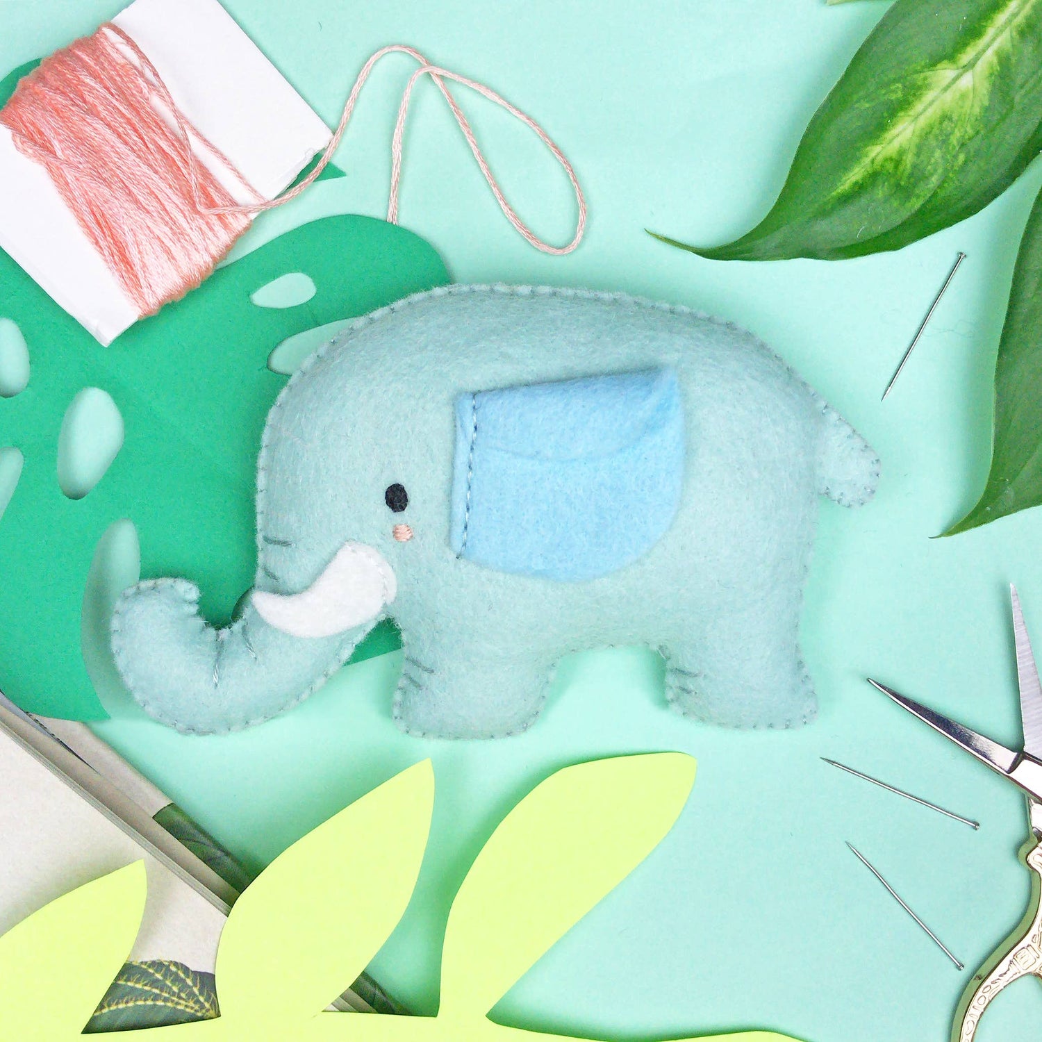 En liten blå elefant i ullfilt ligger tillsammans med en rosa tråd oh grönt blad. 