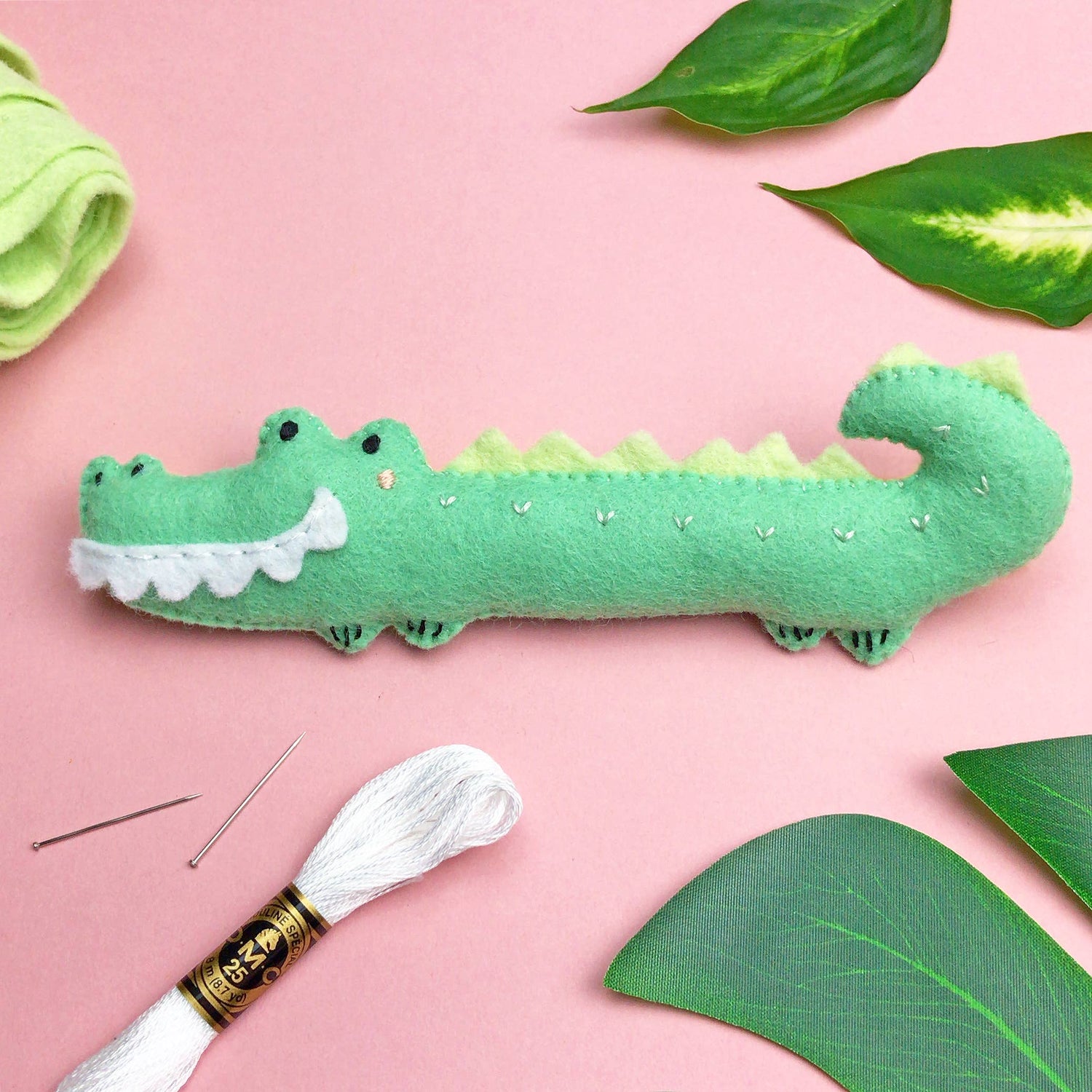 En liten grön krokodil i ekologisk ullfilt ligge spå en rosa plan yta tillsammans med gröna blad. 