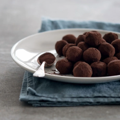 En blå kökshandduk linne som ligger vikt på ett grått bord. På kökshandduken står det en vit tallrik med bruna chokladtryfflar. På tallriken ligger en vit tesked med kakao pulver på. 