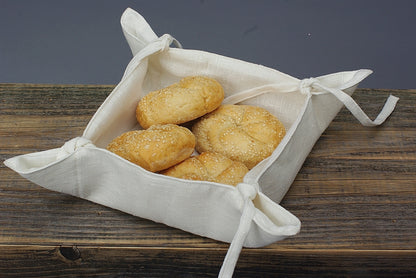 Kvadratisk brödkorg i vit linne med knytband på alla fyra sidor. I brödkorgen ligger 4 stycken bullar. 