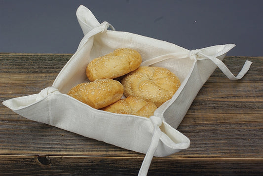 Kvadratisk brödkorg i vit linne med knytband på alla fyra sidor. I brödkorgen ligger 4 stycken bullar. 