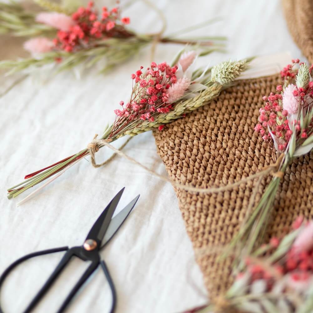 Blombuketter av torkade blommor hänger i ett jutesnöre och ligger på ett bord. 